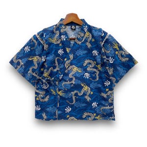 Peut inclure: Chemisier bleu de style kimono avec un motif de dragon. Le chemisier présente un col en V et des manches courtes. Le motif de dragon est en or et blanc, sur un fond bleu avec des motifs de nuages. Le chemisier est présenté sur un cintre en bois.