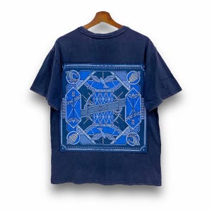 May include: Navy blue t-shirt with a large, detailed blue graphic on the back. The graphic features geometric shapes, birds, and the word "ON" in a banner. The shirt is hanging on a wooden hanger.