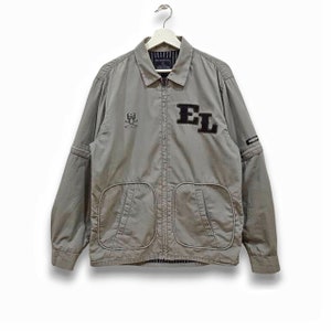 May include: A gray zip-up jacket with a black embroidered skull and crossbones on the left chest and the letters "EL" in black on the right chest. The jacket has two pockets on the front and a black zipper.