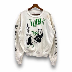 Puede incluir: Sudadera blanca de manga larga con un diseño de panda y bambú. El texto "HONG KONG" está impreso en la parte delantera de la sudadera. Las mangas tienen el texto "STEPHANIE" y "中文排版" impreso en ellas.