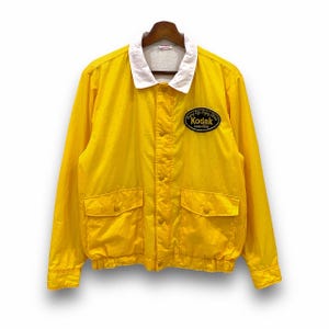 May include: A yellow jacket with a white collar and a black Kodak logo patch. The jacket has two pockets and a zipper closure.