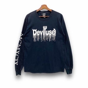 May include: Black long-sleeve shirt with the white text "Deviluse" in a dripping font on the front. The left sleeve has the white text "MCMXCIX". The shirt is hanging on a wooden hanger.
