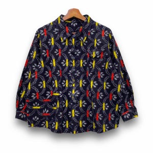 Puede incluir: Camisa azul marino con un estampado repetido de líneas verticales amarillas, rojas y grises y formas florales. La camisa de manga larga tiene cuello y dos bolsillos en el pecho. La camisa está colgada en una percha de madera.