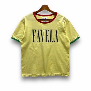 Puede incluir: Camiseta amarilla con la palabra "FAVELA" impresa en negro. La camiseta tiene cuello rojo y ribete verde en las mangas. La camiseta está colgada en una percha de madera.
