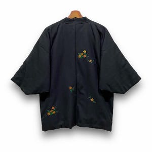 Könnte beinhalten: Schwarze Kimono-Jacke mit weiten Ärmeln und lockerer Passform. Die Jacke hat gestickte Blumenmuster in Orange- und Gelbtönen. Das Kleidungsstück wird auf einem Holzbügel vor weißem Hintergrund präsentiert.
