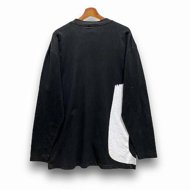 Puede incluir: Camiseta negra de manga larga con un dise&ntilde;o abstracto blanco en el lateral. La camiseta est&aacute; colgada de una percha de madera sobre un fondo blanco. El dise&ntilde;o parece ser de un material reflectante texturizado.