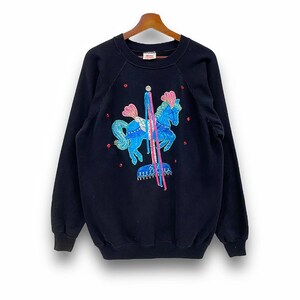 May include: Black sweatshirt featuring a carousel horse design. The horse is blue with pink accents and a silver saddle. The design includes red and silver embellishments. The sweatshirt has a crew neck and long sleeves.