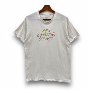 Pode incluir: T-shirt branca com a frase "REX ORANGE COUNTY" em letras coloridas de arco-íris, em estilo manuscrito. A t-shirt está pendurada num cabide de madeira. Tem mangas curtas e gola redonda.