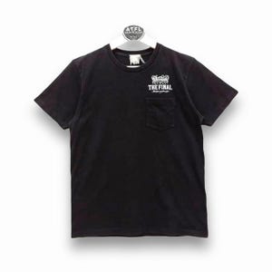Op de afbeelding: Zwart T-shirt met een zakje op de voorkant. Het zakje heeft een witte afbeelding met de tekst "The Final" met een schedel en gekruiste beenderen.