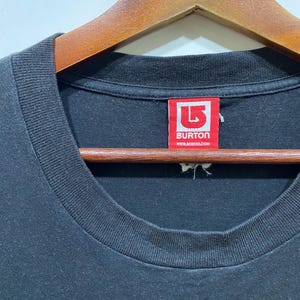 Puede incluir: Primer plano de una camiseta negra con cuello redondo. Una etiqueta roja con el texto blanco "BURTON" y "www.burton.com" es visible. La camiseta est&aacute; colgada en una percha de madera.