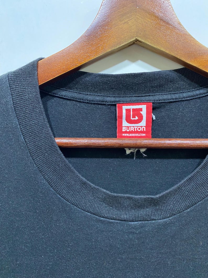 Puede incluir: Primer plano de una camiseta negra con cuello redondo. Una etiqueta roja con el texto blanco "BURTON" y "www.burton.com" es visible. La camiseta est&aacute; colgada en una percha de madera.