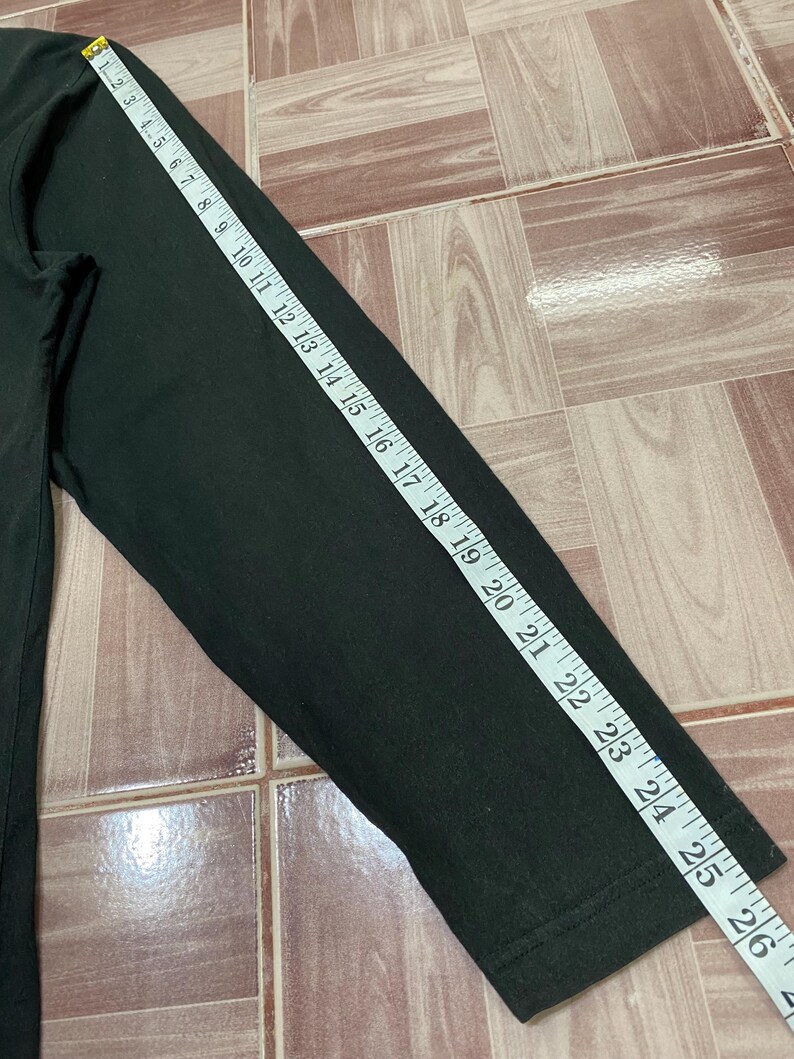 Puede incluir: Camiseta negra de manga larga, con una manga que mide 66 cm. La camiseta es de tela oscura y se mide con una cinta m&eacute;trica blanca. La camiseta est&aacute; sobre una superficie embaldosada.