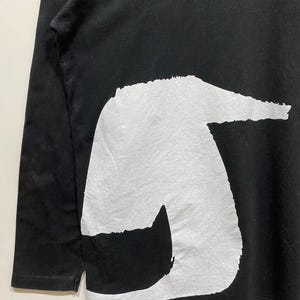 Puede incluir: Camiseta negra de manga larga con un gran dise&ntilde;o abstracto blanco en la parte delantera. El dise&ntilde;o tiene un aspecto texturizado. Las mangas son largas y la tela parece suave.