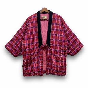 May include: A quilted kimono-style jacket in a red and purple plaid pattern. The jacket features a black velvet collar and trim, a pink lining, and a tie closure. The garment is displayed on a wooden hanger.