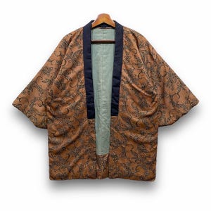 Könnte beinhalten: Braune Jacke im Kimono-Stil mit schwarzem Blumenmuster. Die Jacke hat einen marineblauen Kragen und Besatz sowie ein hellgrünes Innenfutter. Sie hängt an einem Holzkleiderbügel.