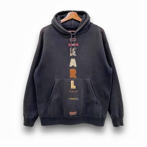 Może przedstawiać: Czarna bluza z kapturem z napisem "KARL" w literach z patchworku z przodu. Bluza ma kieszeń typu kangur i kaptur ze sznurkiem. Marka "Helmut" jest wydrukowana poniżej.