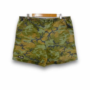 Könnte beinhalten: Tarnshorts in Grün-, Braun- und Schwarztönen mit elastischem Bund und Taschen. Die Shorts tragen den Text "US ARMY" auf der unteren linken Seite. Die Shorts hängen an einem Holzbügel.