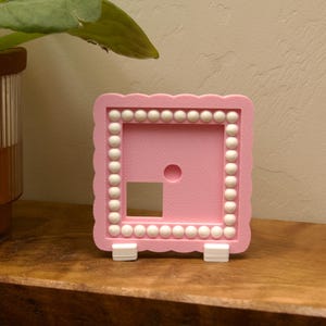 May include: A square pink frame with a scalloped edge and a border of white spheres. The frame features a central circular cutout and a square cutout in the lower left corner. It is supported by two small white stands at the base.