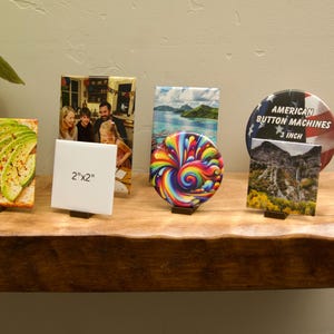 May include: A collection of display items including square tiles with avocado and family photo prints, a white tile labeled 2"x2", a circular button with a rainbow swirl, a landscape tile, and a 3-inch American Button Machines pin with a flag design.