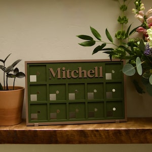 May include: A rectangular olive green wooden display board featuring a grid of small square cutouts and the name Mitchell in raised brown lettering. The frame is a muted brown color, and the board is designed to stand upright on a flat surface.