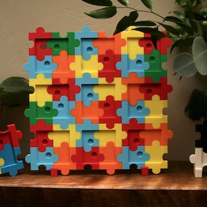May include: A large square wall display board composed of interlocking puzzle-shaped pieces in red, orange, yellow, blue, and green, featuring nine square cutouts. Two smaller, separate puzzle-shaped display pieces are positioned on either side.