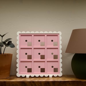 Pode incluir: Um organizador de exposição quadrado cor-de-rosa com nove compartimentos individuais, cada um com um pequeno orifício circular e um recorte quadrado. O organizador é emoldurado por uma borda branca recortada.