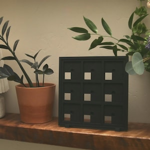 May include: A black square display frame featuring a grid of nine individual compartments, each with a small circular opening. The frame is constructed with a solid border and thin dividers, creating a uniform arrangement of nine square-shaped slots.