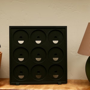 May include: A square black display rack featuring a grid of nine circular cutouts arranged in three rows and three columns. Each circular opening contains a small central hole and a semi-circular cutout at the bottom edge of the inner circle.