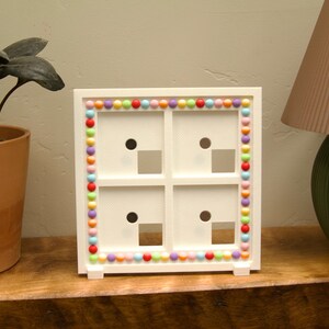 May include: A square white frame with four equal compartments, each featuring a circular hole and a square cutout. The outer border of the frame is decorated with a continuous row of small, multicoloured beads in shades of red, blue, yellow, and pink.