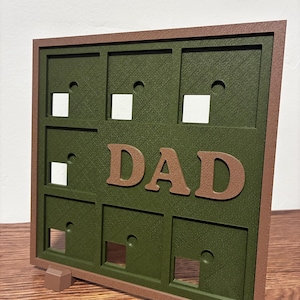 Könnte beinhalten: Ein quadratisches, olivgrünes, 3D-gedrucktes Anzeigetafel-Objekt mit braunem Rahmen und Sockel. Die Tafel verfügt über ein Raster aus neun vertieften Quadraten, die jeweils eine kleine kreisförmige Vertiefung aufweisen. Das Wort DAD steht mittig.