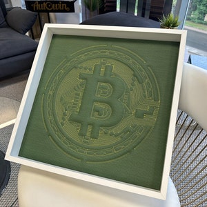 May include: A framed green fabric artwork with a yellow embroidered Bitcoin symbol. The symbol is surrounded by concentric circles.