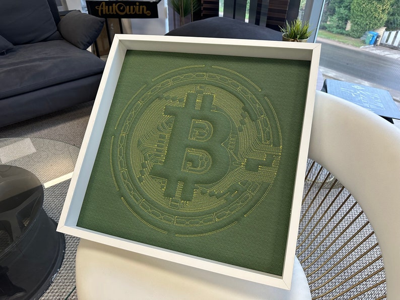 May include: A framed green fabric artwork with a yellow embroidered Bitcoin symbol. The symbol is surrounded by concentric circles.