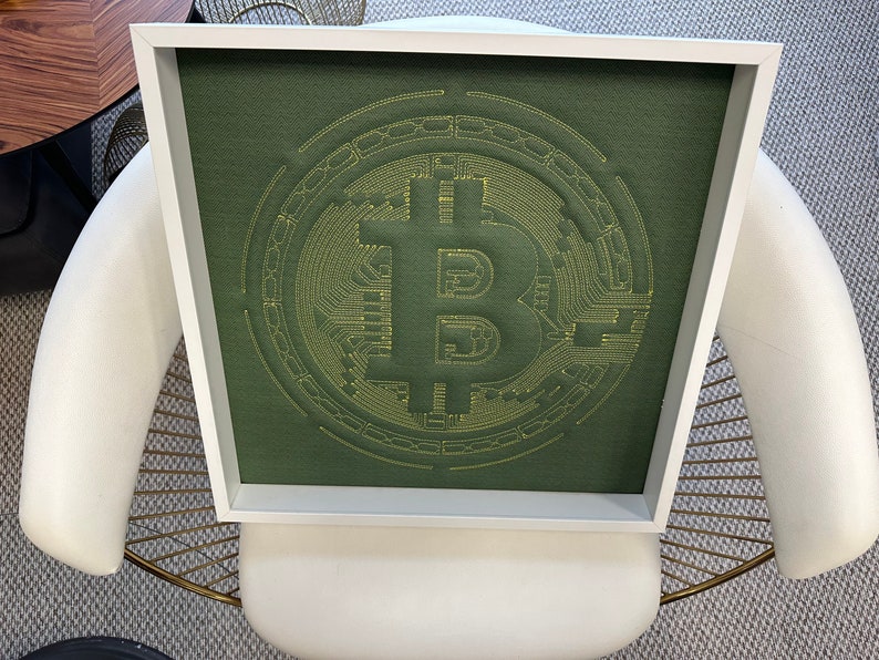 May include: A framed green canvas with a yellow embroidered Bitcoin logo. The logo is a stylized letter B with circuit board designs.