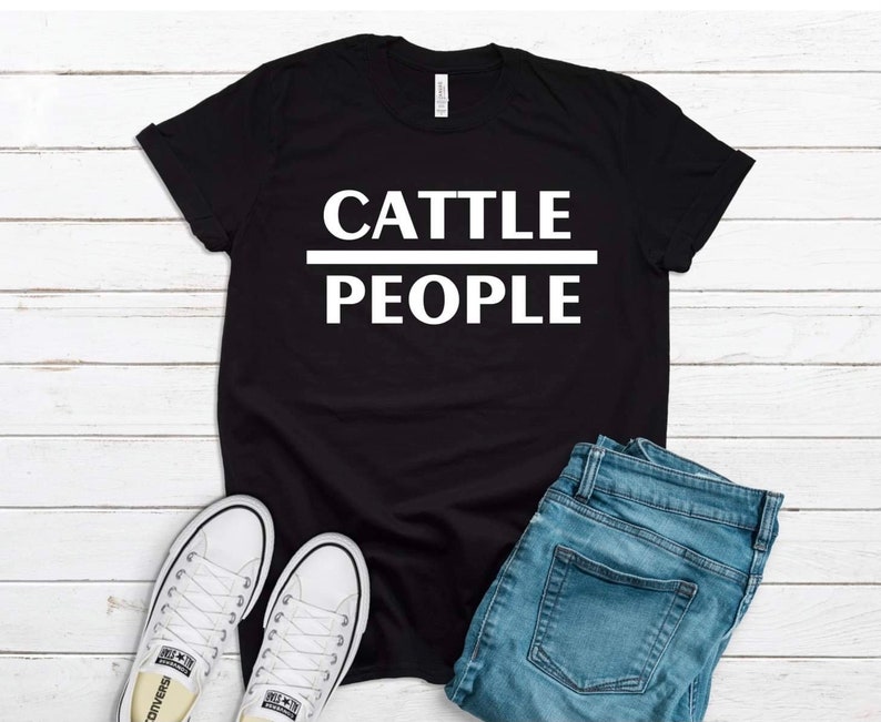 May include: Black t-shirt with white text that reads "Cattle People". The shirt is laid flat on a white wooden surface with a pair of white sneakers and blue jeans.