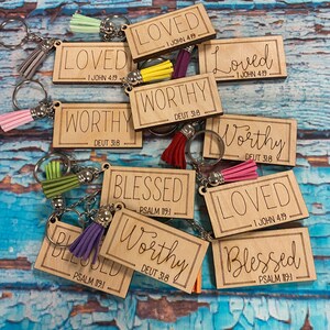 May include: A set of wooden keychains with different inspirational words and bible verses. The keychains are decorated with colorful tassels. The words on the keychains include "Loved", "Worthy", and "Blessed".