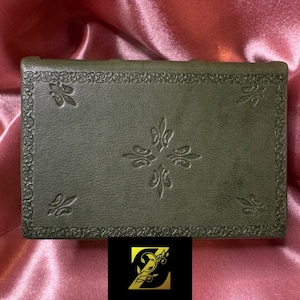 May include: A dark green leather-bound journal with an intricate embossed design. The cover features a repeating pattern of stylized fleur-de-lis.