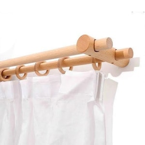 Puede incluir: Un juego de barras de cortina dobles de madera con cortinas blancas. Las barras son de color marrón claro y cuentan con anillos de madera para colgar las cortinas. Las cortinas son blancas y parecen estar hechas de una tela ligera. Las barras están montadas en una pared blanca.