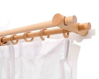 May include: A double curtain rod system made of light-colored wood, featuring two parallel horizontal bars supported by a wall-mounted bracket. Several wooden rings are attached to the lower rod, holding a white, pleated fabric curtain.