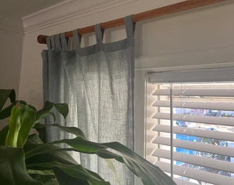 May include: A light blue curtain panel with a tab top header hangs from a wooden curtain rod. The fabric has a visible woven texture and is positioned over a window with white horizontal blinds. A green plant is visible in the foreground.