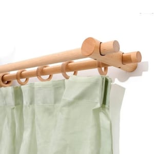 Puede incluir: Un sistema de barra de cortina doble hecho de madera de tono claro, con dos barras horizontales sostenidas por soportes de pared. Varios anillos circulares de madera están unidos a la barra inferior, sosteniendo una cortina de tela verde salvia.