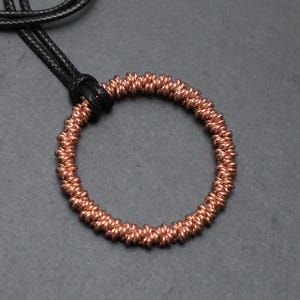 May include: A circular pendant crafted from coiled copper wire, featuring a textured, twisted metal surface. The pendant is suspended from a simple black cord, creating a contrast between the metallic copper tones and the dark cord material.