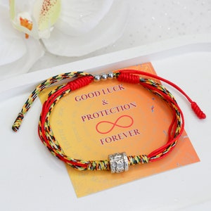 May include: Red and yellow braided string bracelet with silver beads and a charm that says "GOOD LUCK & PROTECTION FOREVER" on a white background.