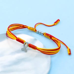 May include: A red and yellow cord bracelet with a silver ribbon charm. The bracelet features adjustable sliding knots and small silver beads. The cord is a combination of red and yellow.