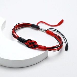 May include: Red and black braided bracelet with a knot design and silver beads. The bracelet has a tassel on each end.