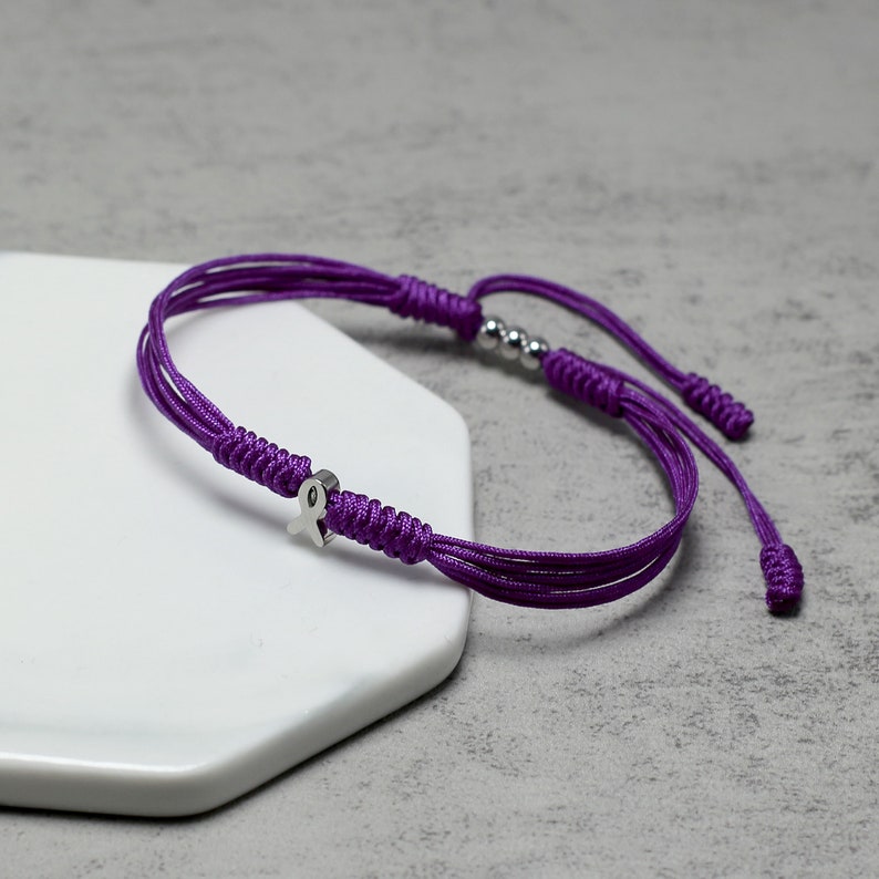 K&ouml;nnte beinhalten: Lilafarbenes geflochtenes Armband mit einem silbernen Schleifenanh&auml;nger. Das Armband hat verstellbare Schiebeknoten.