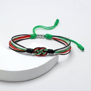 May include: A green, white, red, and black braided bracelet with a knotted design. The bracelet has a silver bead accent.