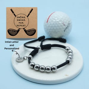May include: A black cord bracelet with silver beads and a personalized initial charm. A golf ball and a card with golf club graphics and the text "SWING. SWEAR. PAR. REPEAT." are also in the image.