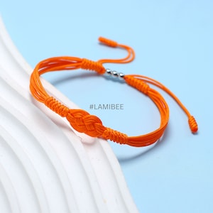 May include: Orange braided friendship bracelet with a silver bead accent and a knot in the center. The bracelet has two long strands that can be adjusted for a custom fit. #LAMIBEE