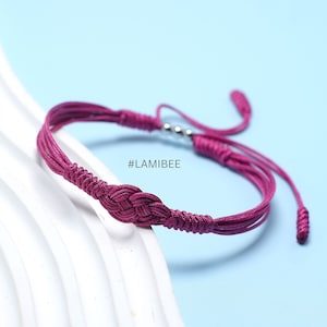 Könnte beinhalten: Ein geflochtenes Armband in Lila mit einer silbernen Perle und einem Knoten in der Mitte. Das Armband hat an jedem Ende einen Quasten. #LAMIBEE