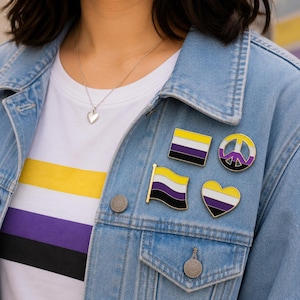 May include: A light blue denim jacket worn over a white t-shirt featuring a yellow, white, purple, and black horizontal striped pattern. Four enamel pins are attached to the jacket, displaying the same yellow, white, purple, and black colors.